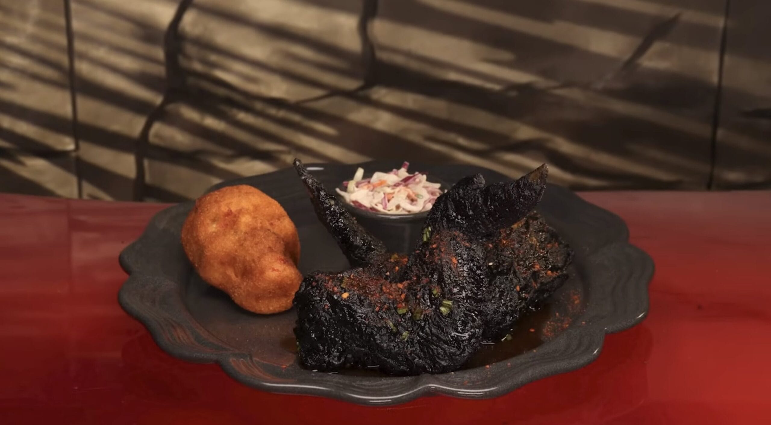 A blackened chicken wing with visible spices, a side of coleslaw in a small bowl, and a piece of fried cornbread await on a dark plate set against a red surface—a vibrant display in the epic universe of food and beverage.
